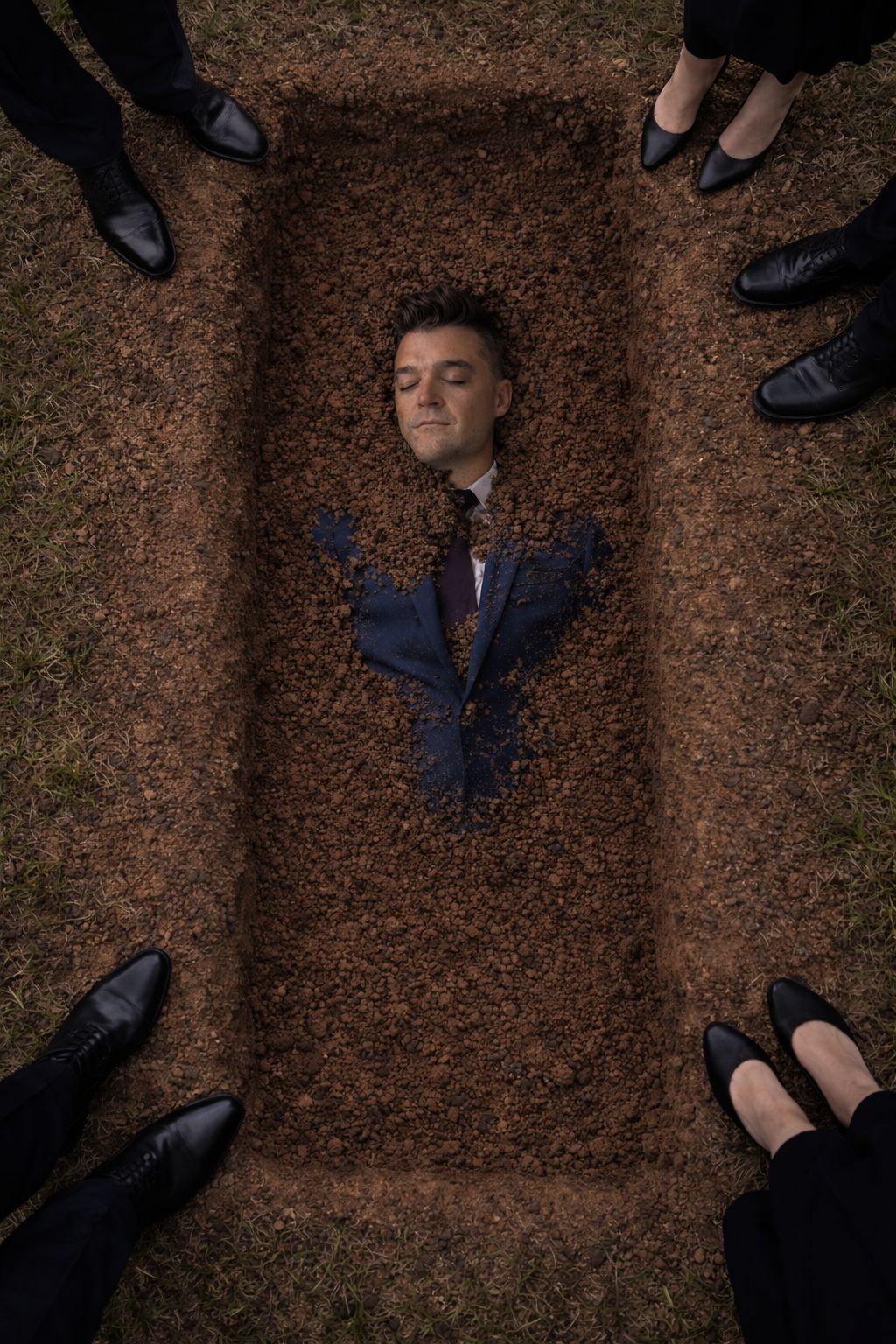 Overhead view of Nathan Morris lying in an open grave, covered in soil to the chest, with mourners' black shoes standing around the edge.