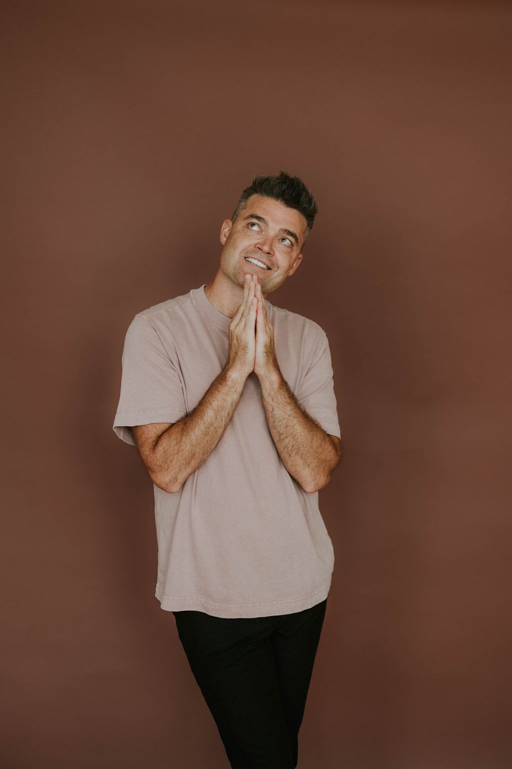 Nathan Morris smiling and looking up, hands pressed together, against a warm brown wall.